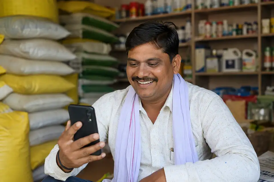 A feed dealer smiling at his phone in his shop, with stacked cattle feed bags and supplement bottles behind him