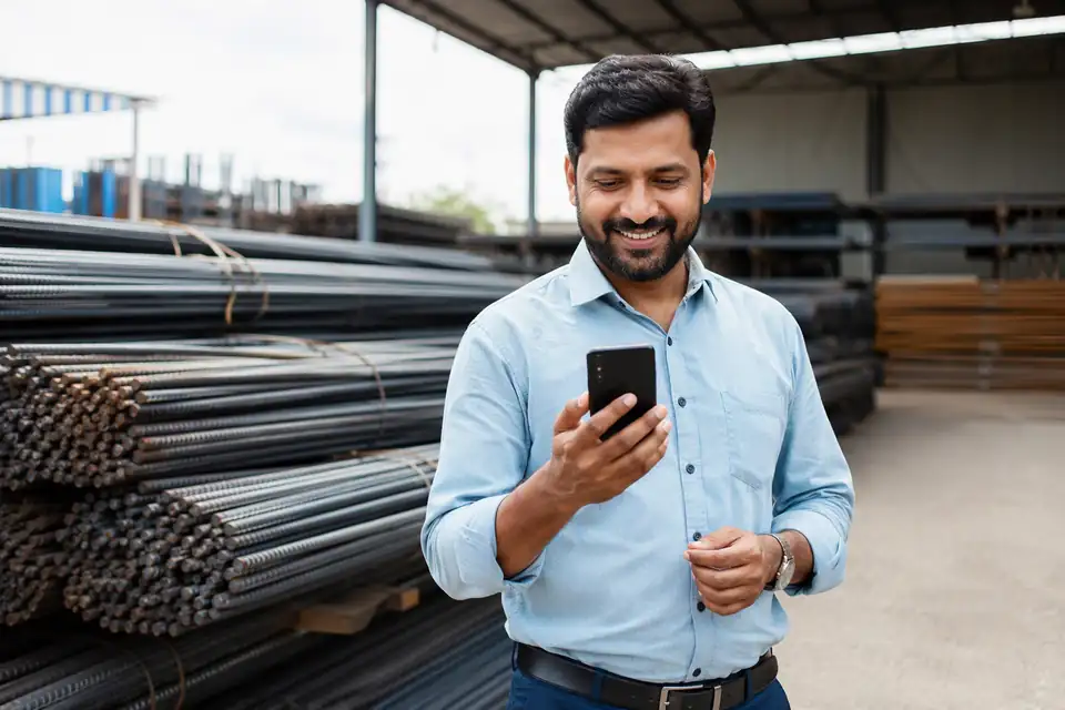 A steel dealer in his yard beside stacks of TMT bars, smiling at his phone