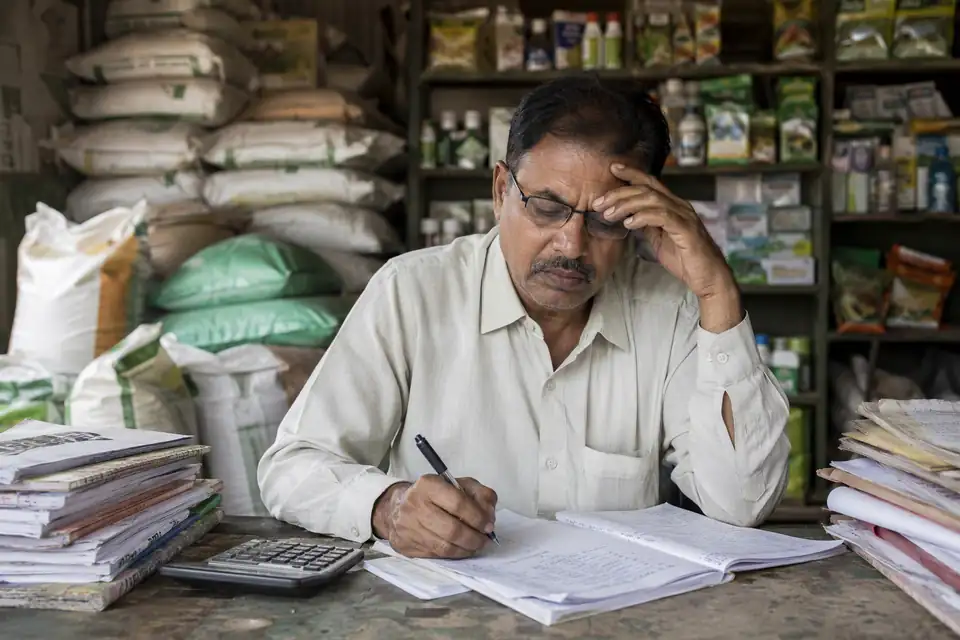 An agro-dealer at his desk working through ledgers and invoices by hand, with fertilizer bags stacked behind him