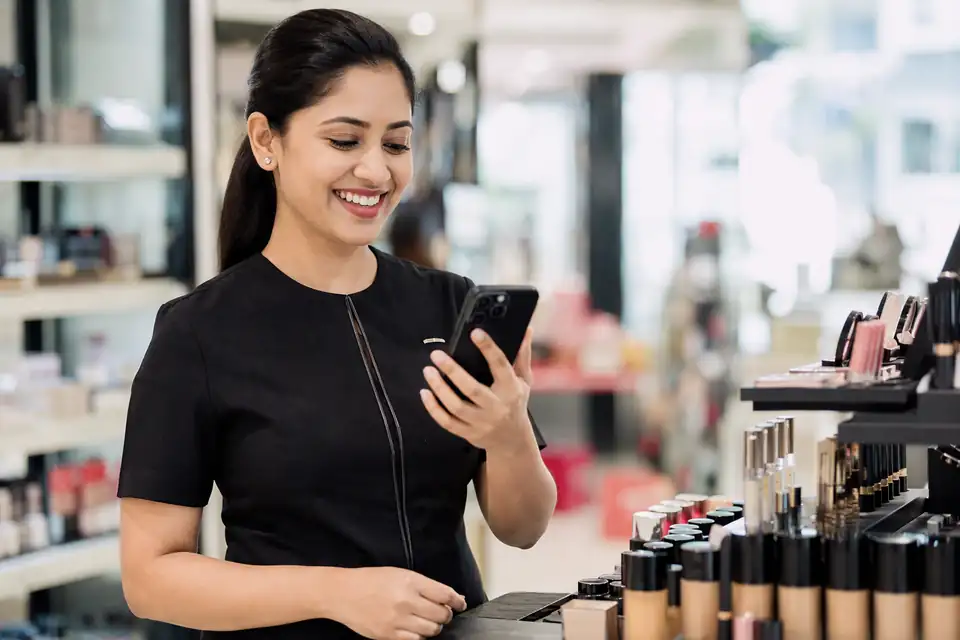 A beauty advisor in a black uniform smiling at her phone beside a makeup display counter