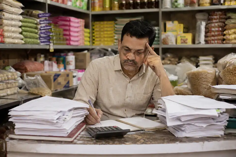 A kirana shop owner frowning over piles of paper bills and a calculator at his counter