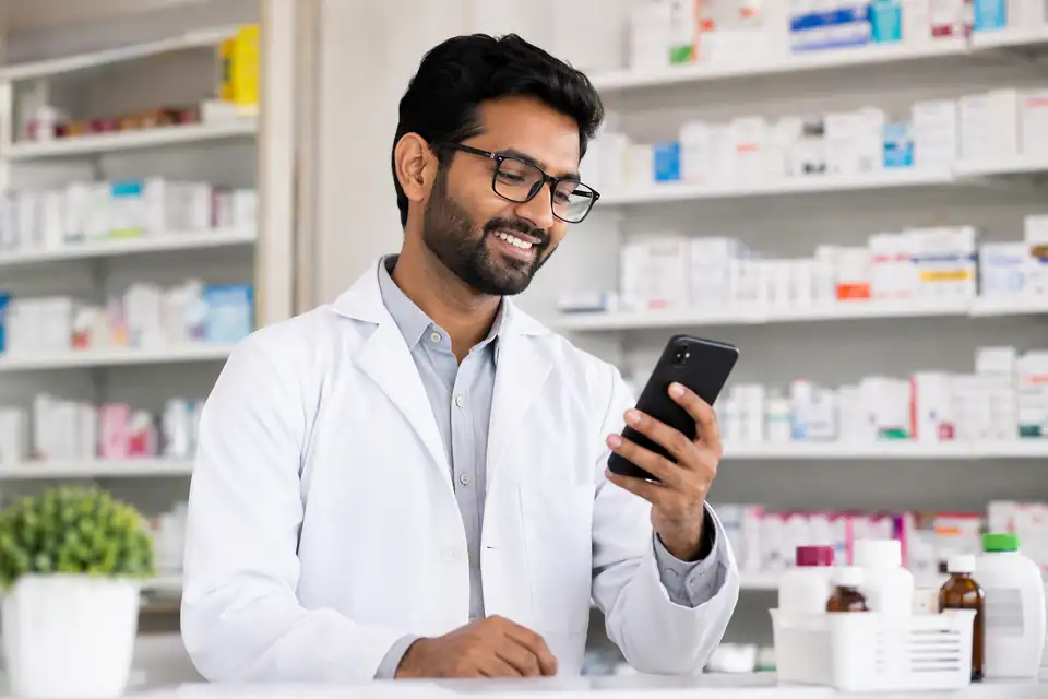 A pharmacist in a white coat smiling at his phone at the counter, with medicine shelves behind him